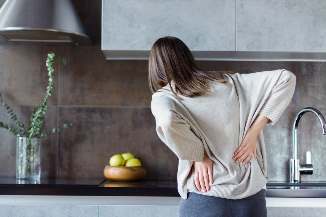 Woman bending forward holding her lower back with both hands in kitchen showing pain and fatigue from prolonged standing on hard floors