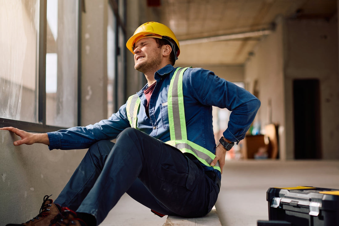Construction worker in hard hat and safety vest sitting on floor, holding lower back in pain.