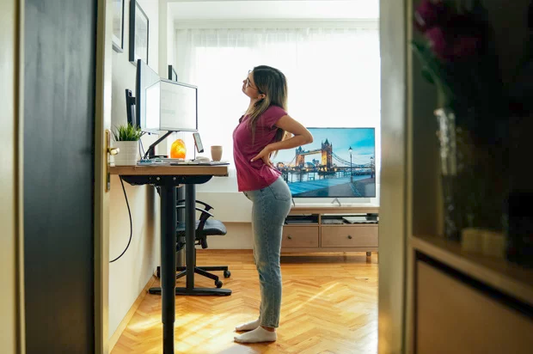 Woman standing barefoot at a standing desk holding her lower back in pain on hard wood floor without anti-fatigue mat