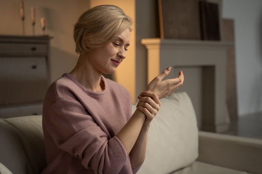 Middle-aged woman in pink sweater sitting on sofa holding her wrist with a pained expression showing wrist pain from arthritis, carpal tunnel, or repetitive strain injury