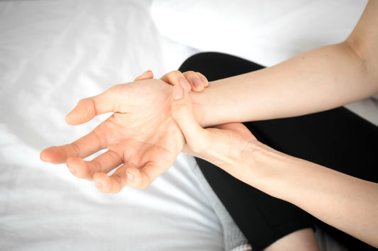 Woman sitting on bed in white shirt holding her wrist with other hand showing nighttime wrist pain from carpal tunnel syndrome, tingling, or numbness during sleep