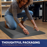 Woman in striped shirt kneeling and smoothing out gray FEATOL anti-fatigue standing mat on office floor, demonstrating thoughtful non-folding roll packaging for easy unboxing and immediate use